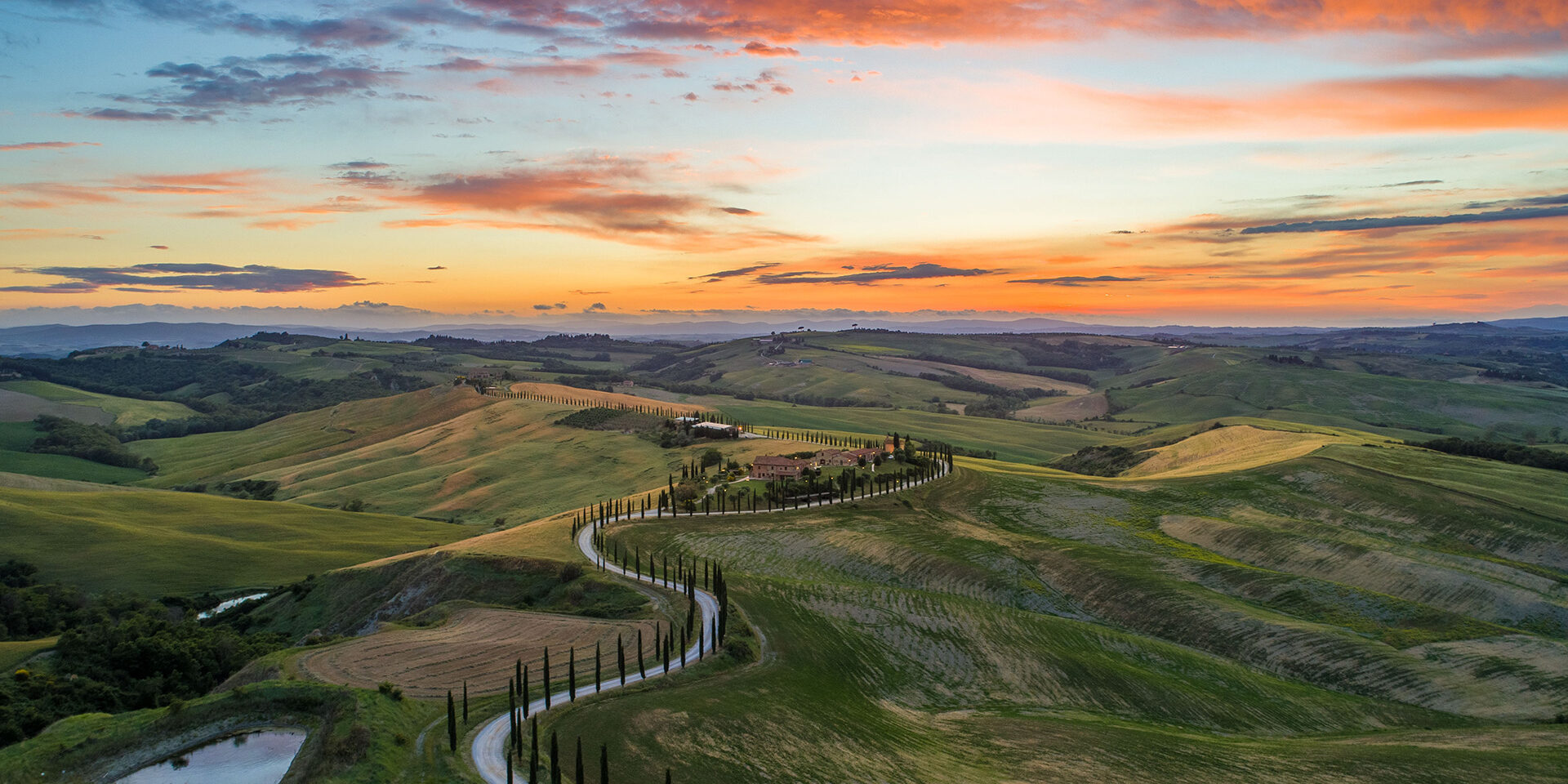 road-between-green-grass-field-near-mountains-under-blue-and-brown-sky-at-golden-hour